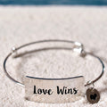 A silver-toned metal bangle bracelet with a rectangular plate engraved 'Love Wins' and a small heart charm, resting on a textured white surface.