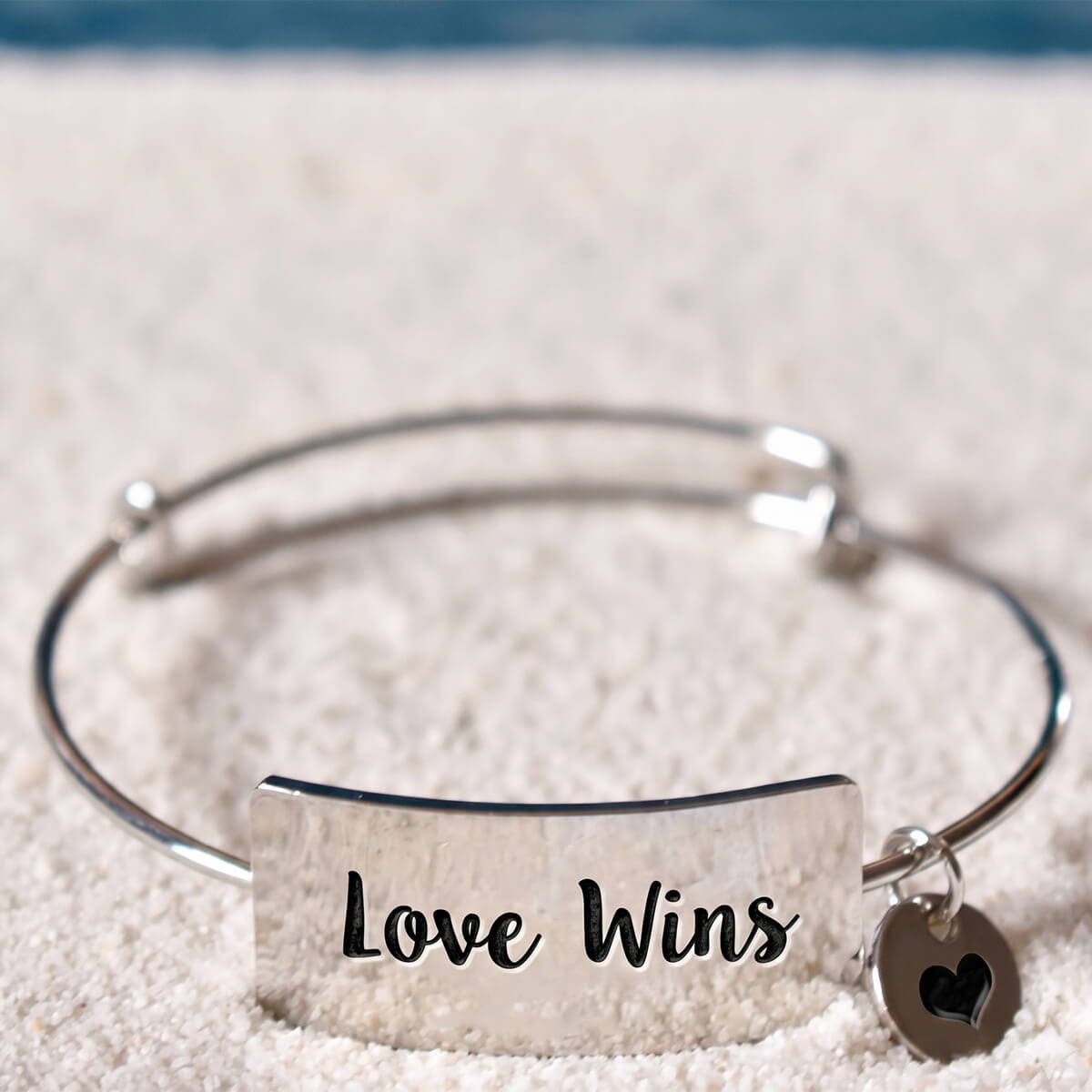 A silver-toned metal bangle bracelet with a rectangular plate engraved 'Love Wins' and a small heart charm, resting on a textured white surface.